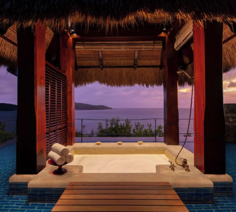A luxurious outdoor bathtub under a thatched roof gazebo overlooks the ocean at sunset, surrounded by blue tiles and wooden beams, with rolled towels neatly placed nearby.