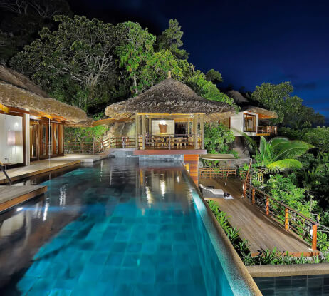 Luxurious tropical villa with thatched roofs, surrounded by lush greenery. An illuminated infinity pool stretches alongside wooden decks at night, reflecting the lights from the open, modern living spaces.
