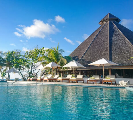 A luxurious resort with a thatched-roof pavilion, poolside lounge chairs under white umbrellas, palm trees, and a clear blue swimming pool under a sunny sky.