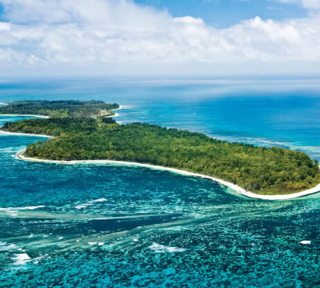 Aerial view of a lush green island surrounded by turquoise blue ocean waters and white sandy beaches, with waves breaking along the shoreline under a partly cloudy sky.