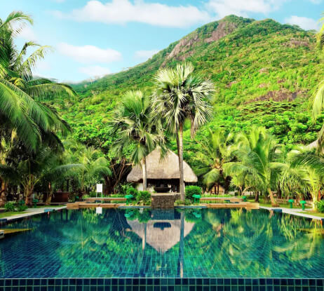 A tropical resort pool surrounded by palm trees, lush greenery, and mountains in the background under a blue sky with scattered clouds. A thatched-roof hut sits beyond the pool.