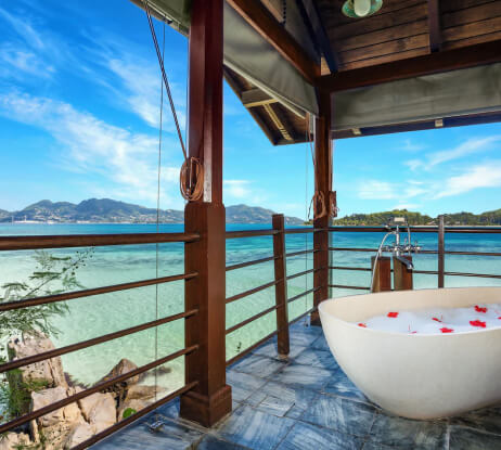 A private outdoor bathtub filled with water and rose petals sits on a terrace overlooking a clear blue ocean and distant mountains under a bright sky.