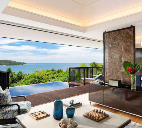 A luxurious living room opens to a wooden deck with an infinity pool, overlooking lush greenery and a blue ocean under a bright sky. The room features modern decor, a chessboard, and tropical plants.