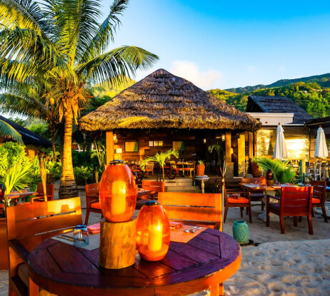 Outdoor beach restaurant with wooden tables and chairs, orange lanterns, palm trees, and a thatched-roof hut, bathed in warm sunlight with mountains in the background.