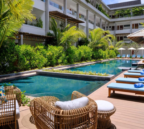 Outdoor pool area at a modern hotel, featuring wicker chairs, blue cushioned loungers, palm trees, lush greenery, and a wooden deck beside a clear rectangular swimming pool.