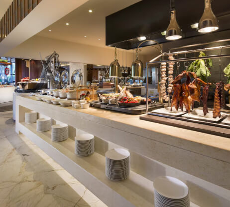 A hotel buffet features various hot dishes, salads, and roasted meats hanging on display. Plates are neatly stacked below the counter on marble flooring in a bright, modern dining area.