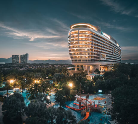 A large, modern hotel with a unique, curved design is illuminated at dusk, overlooking a lush, tropical area with a brightly lit outdoor pool and water play area, surrounded by trees and distant buildings.