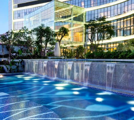 A modern glass building with illuminated windows stands behind a decorative fountain with cascading water and blue-lit pool, surrounded by lush greenery and trees at dusk.