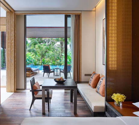 Modern hotel room with a table, chairs, and sofa, decorated with orange pillows and a bowl of fruit. Large glass doors open to a patio with lounge chairs and a pool, surrounded by lush greenery.