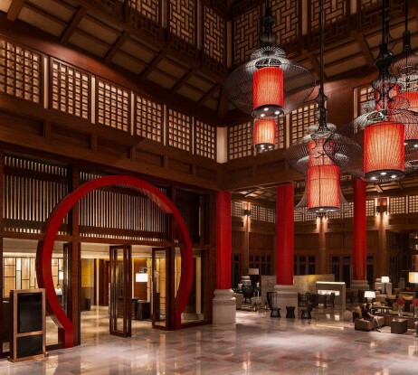Spacious hotel lobby with a high wooden ceiling, large red columns, intricate lattice windows, red circular doorway, and ornate hanging red lanterns illuminating seating areas.