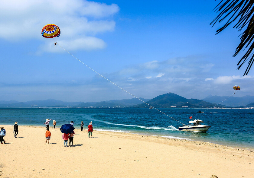 People on a sandy beach watch as a speedboat pulls someone parasailing over the blue water, with mountains and clouds in the background. Some people stand under umbrellas while others walk along the shore.