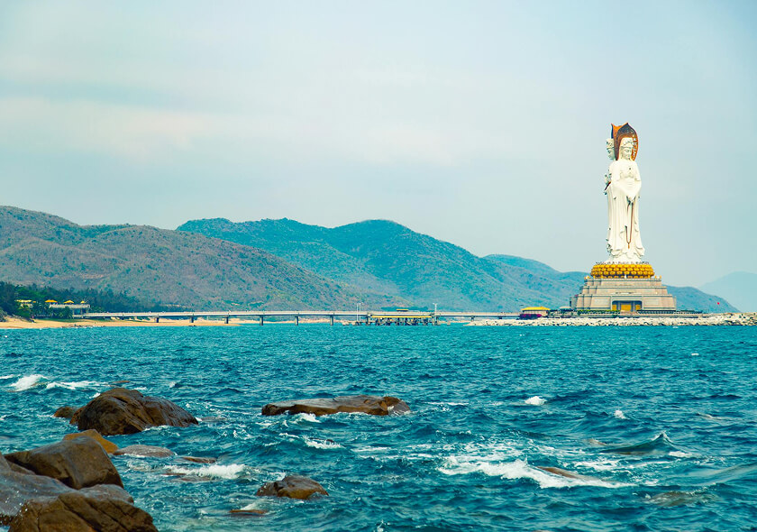 A large white statue stands on a platform by the shoreline, with blue ocean waves in the foreground and distant, tree-covered mountains under a cloudy sky in the background.