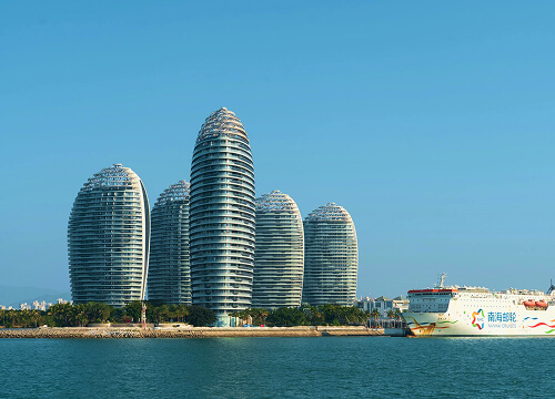 Five modern, oval-shaped high-rise buildings stand along the waterfront under a clear blue sky, with a large white ferry docked nearby. The scene is calm, with water in the foreground.