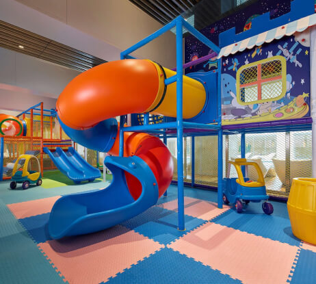 Indoor childrens play area with a blue, red, and yellow spiral slide, toy cars, small slides, and colorful padded flooring. The walls are decorated with playful, cartoon-like space and house designs.