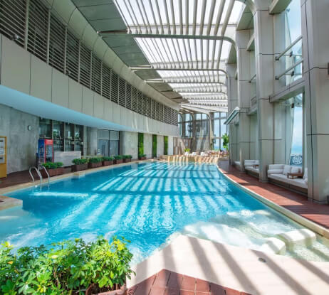 A modern indoor swimming pool with clear blue water, surrounded by lounge chairs and potted plants, under a large open slatted roof letting sunlight stream in.