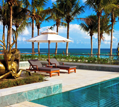 Two lounge chairs sit under a white umbrella beside a pool, with palm trees, a bronze statue, and the ocean visible in the background on a sunny day.