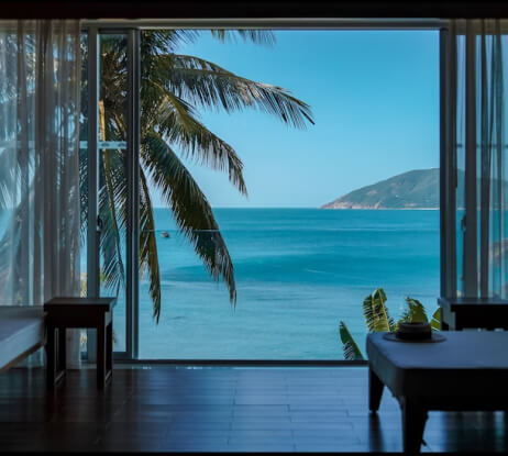 A scenic ocean view seen through large glass doors, with palm trees framing the turquoise water and a distant island. Inside, there are silhouettes of furniture, creating a peaceful and inviting atmosphere.