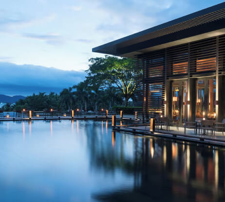 Modern building with large glass windows beside a calm reflecting pool at dusk, surrounded by palm trees and softly glowing lights, creating a peaceful and luxurious atmosphere.