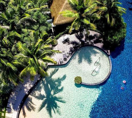 Aerial view of a tropical resort pool surrounded by lush palm trees, wooden deck with lounge chairs, and a thatched-roof building. The pool has clear, shallow and deep blue sections with two submerged lounge chairs.