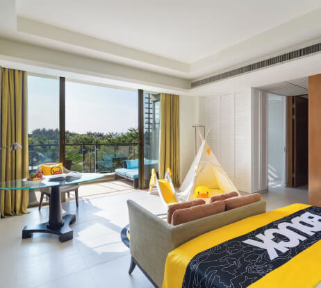 A bright hotel room with large windows, a balcony, a work desk, a teepee tent, and a bed with a Pikachu-themed blanket. Yellow accents and natural light create a cheerful atmosphere.
