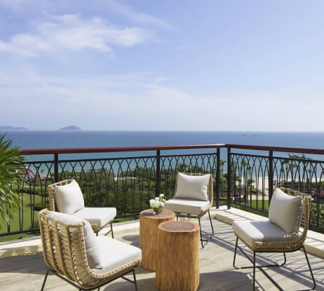 A sunny balcony with four cushioned chairs and two wooden tables overlooks a scenic ocean view with distant islands and ships under a clear blue sky.