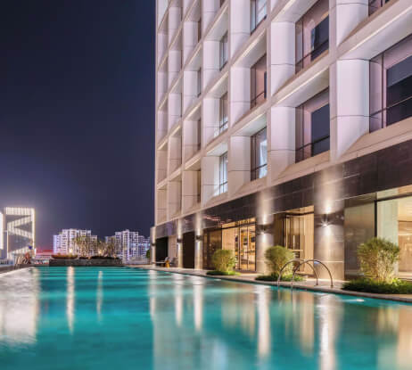 Modern building with large windows beside a well-lit outdoor swimming pool at night; city lights and other buildings are visible in the background.
