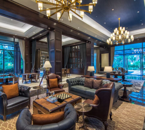Luxurious hotel lounge with leather armchairs, sofas, chandeliers, and large windows overlooking greenery. A chessboard sits on a table, and books are arranged nearby, creating an elegant and inviting atmosphere.