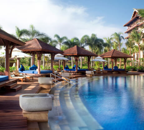 A luxurious outdoor resort pool with curved wooden decking, blue lounge chairs, shaded cabanas, palm trees, and a clear sky in the background.