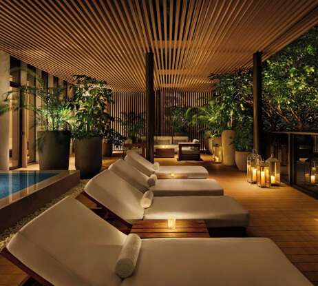 A relaxing spa lounge with cushioned lounge chairs, small tables with candles, potted plants, lanterns, soft lighting, and a tranquil atmosphere near a pool.