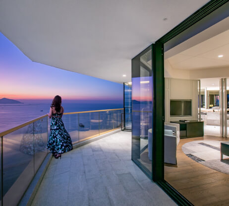 A woman in a long dress stands on a balcony overlooking the ocean at sunset, with a modern living room visible through open glass doors.