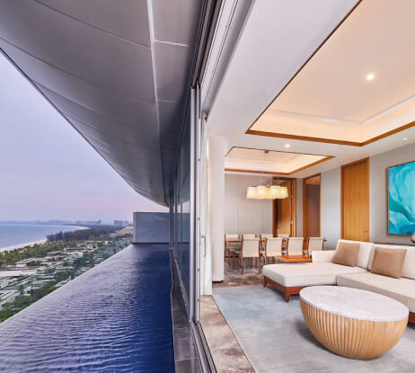 Modern hotel suite with a stylish living area, dining table, and large sliding glass doors opening to a balcony with an infinity pool and ocean view at sunset.