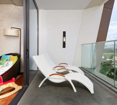 A modern balcony with two white lounge chairs and a glass railing, overlooking a city view. Inside, a cozy seating area with colorful pillows and a childs toy are visible through the open sliding door.