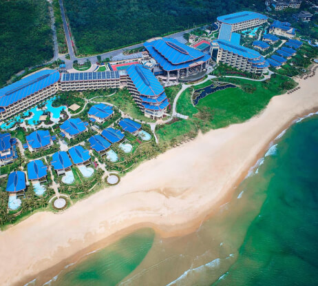 Aerial view of a large beachfront resort with blue-roofed buildings, swimming pools, green gardens, and direct access to a sandy beach and turquoise ocean waves. Dense greenery borders the property.