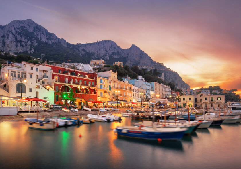 Colorful waterfront buildings and docked boats at sunset in a coastal Italian village, with rocky mountains in the background and soft golden light reflecting on the water.