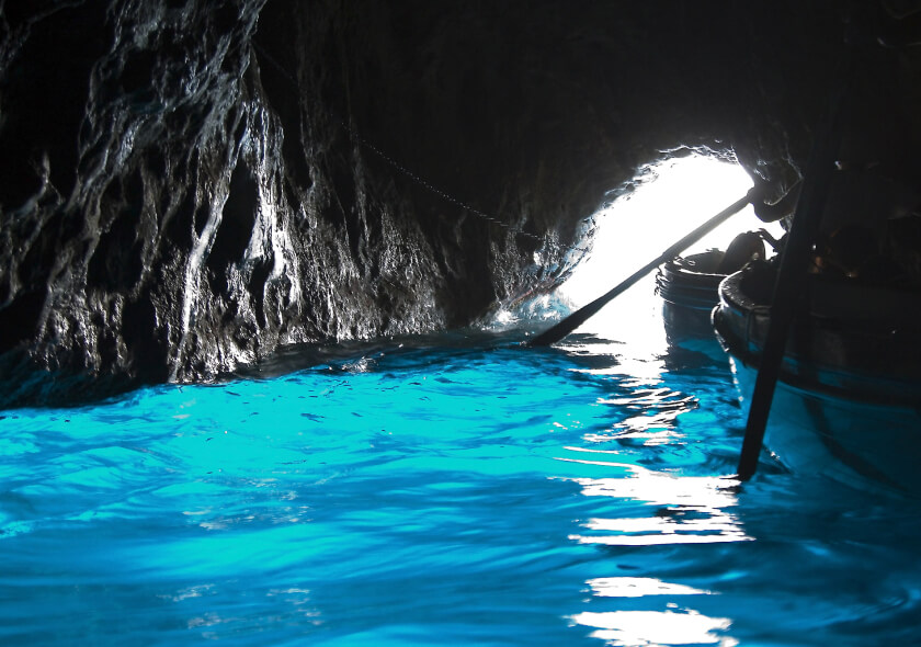 A cave with bright turquoise water reflecting light from an opening. Two boats with oars are partially visible, entering or exiting the cave through the illuminated entrance.