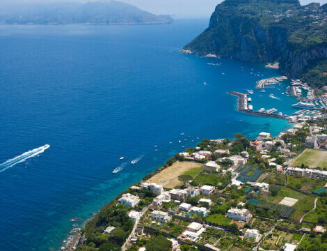 Aerial view of a coastal town with white buildings, green landscapes, and a marina nestled between blue sea and steep cliffs, with boats visible in the water.